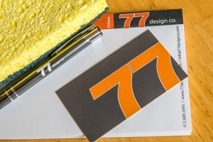 Spring cleaning your business marketing. An image of the 77 Design Co gray and orange business card and notepad along with a yellow and green sponge. 